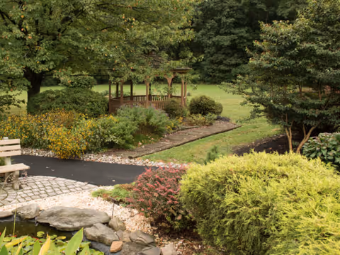 A peaceful outdoor garden area featuring a wooden gazebo surrounded by green trees and bushes, a paved pathway, a small pond with rocks, and a wooden bench.