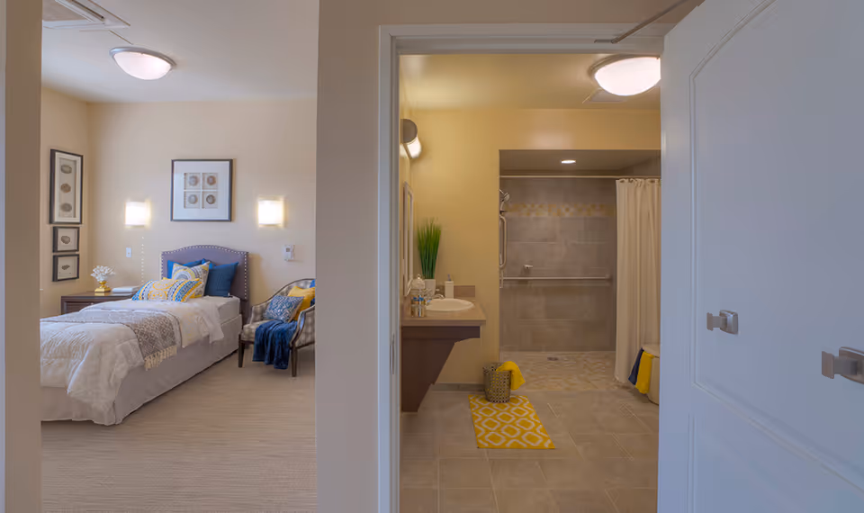 View of a bedroom with a single bed, decorative pillows, a chair with a blanket, and framed artwork on the walls. Adjacent to the bedroom is a bathroom with a sink, a large walk-in shower with a curtain, and a yellow patterned rug on the tiled floor.
