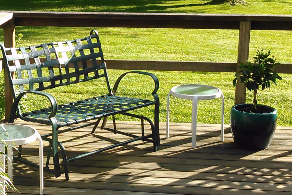 Outdoor wooden deck with a metal bench, two small round tables, and a potted plant. The deck overlooks a green grassy lawn.