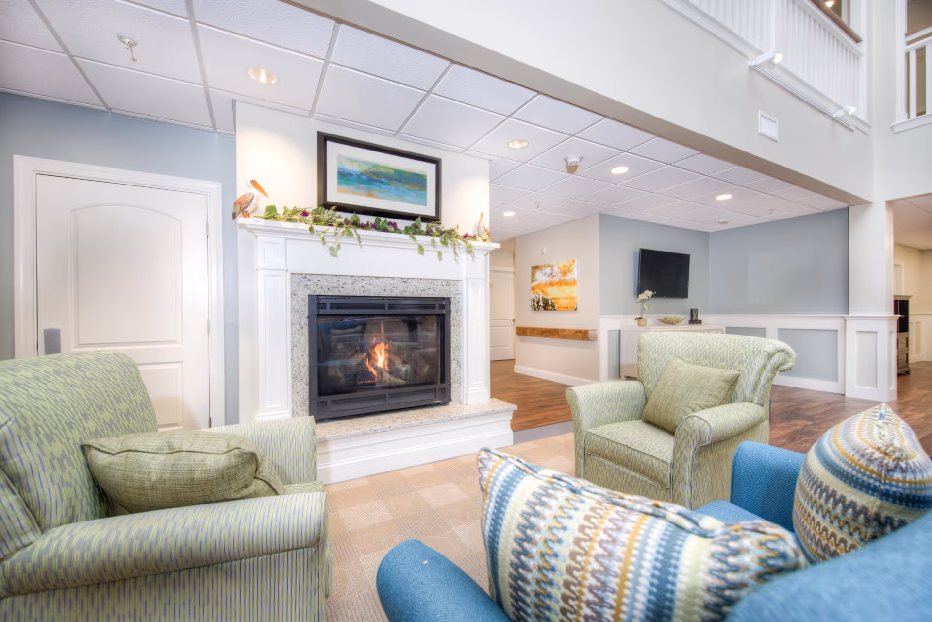 A cozy living room area with a lit fireplace surrounded by a white mantel decorated with greenery and a framed painting above it. There are two green patterned armchairs and a blue chair with a colorful patterned pillow arranged around the fireplace. The room has light blue walls, a white ceiling with recessed lighting, and wood flooring in the background.