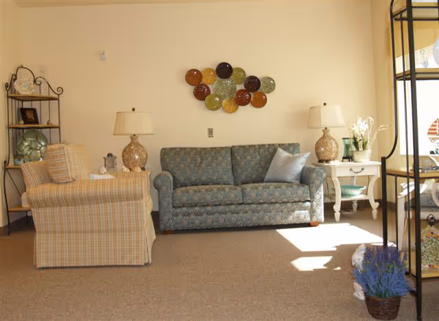 A cozy living room area with a patterned blue sofa, a plaid armchair, two side tables each with a lamp, a decorative metal wall art piece above the sofa, and a shelving unit with decorative items. Sunlight streams in from the right side, illuminating part of the carpeted floor.