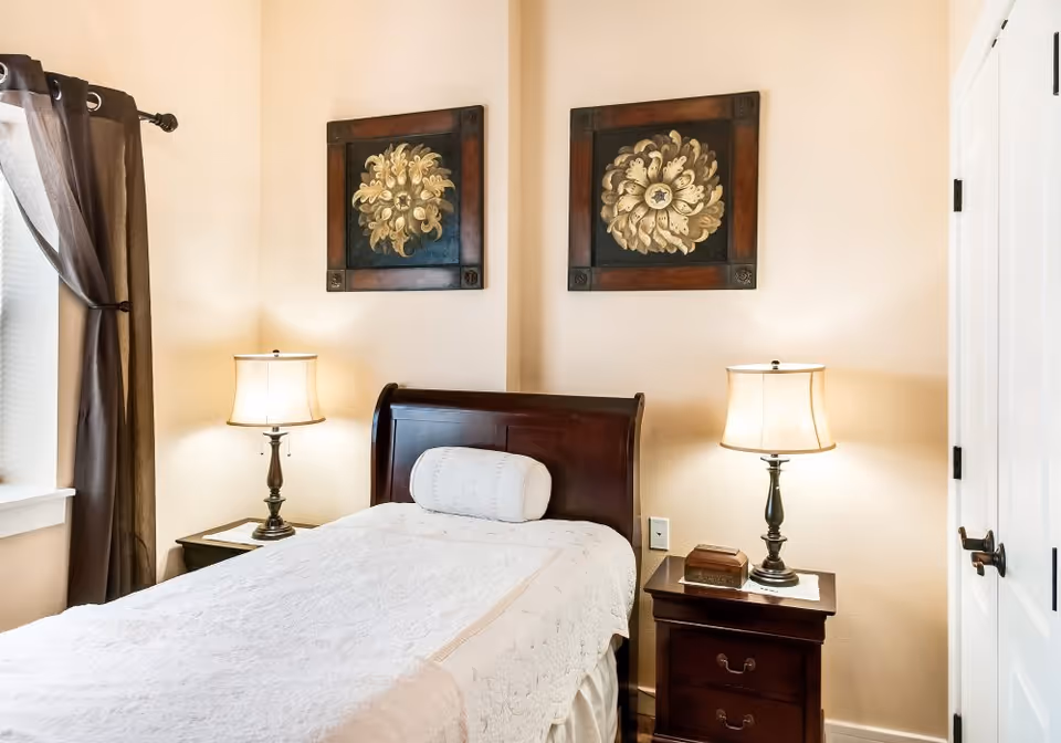 A neatly made single bed in a softly lit bedroom with two bedside tables, lamps, framed wall art, and a curtained window.