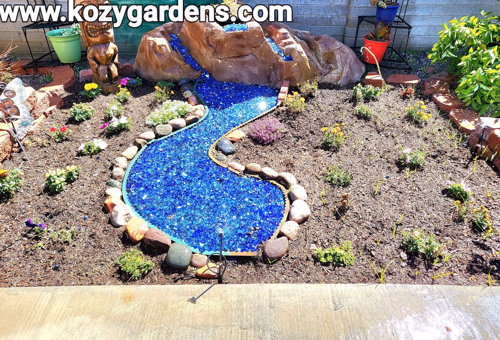 A small garden area with a decorative blue glass stone stream flowing from a rock feature. The garden is bordered with rocks and has various small flowering plants and shrubs planted in mulch. There is a concrete pathway in the foreground and a brick wall in the background. A tiki statue and potted plants are also visible.