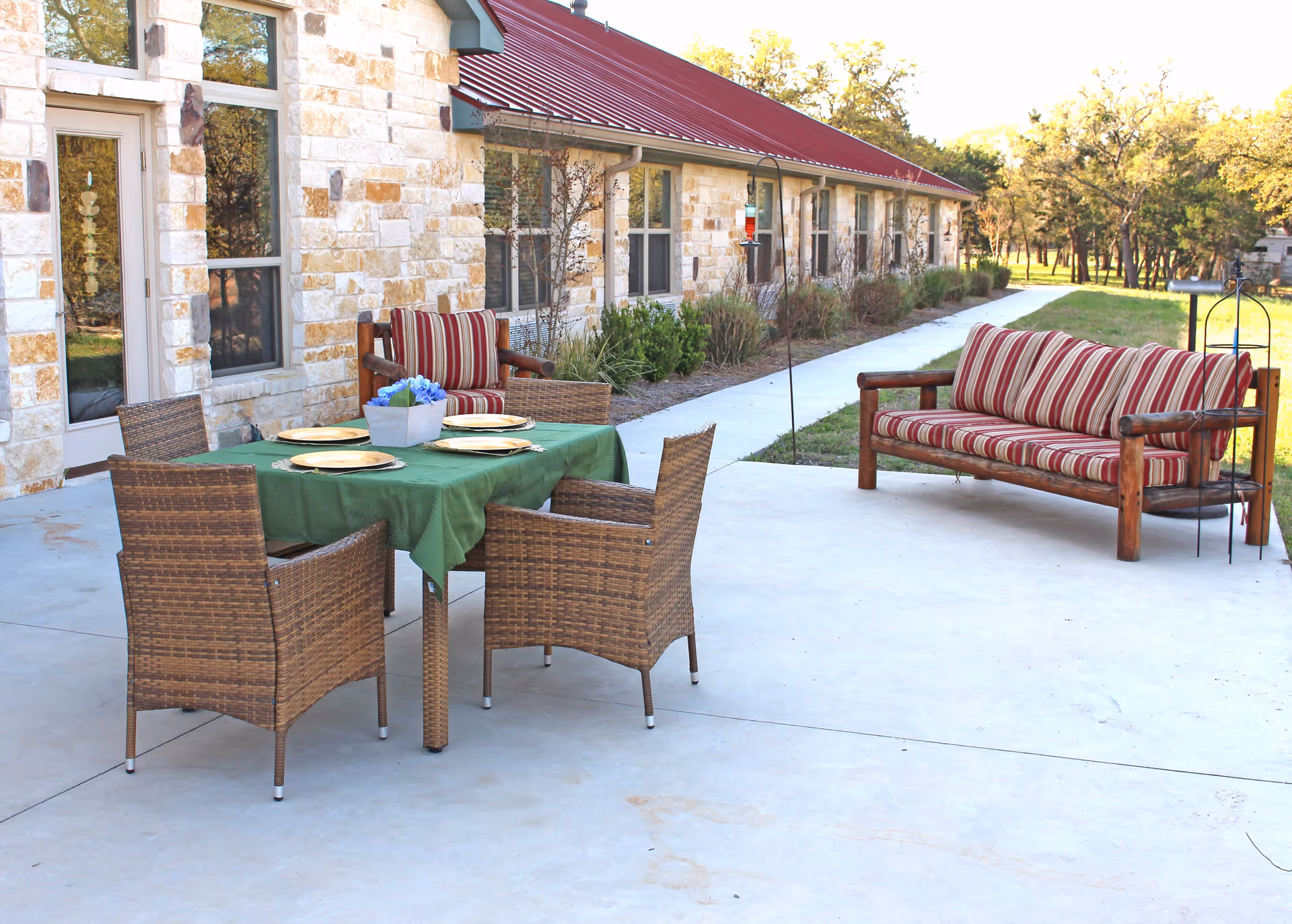 Outdoor patio area at Gemstone Senior Living at Bulverde featuring a stone building with a red metal roof, a table covered with a green tablecloth set with four plates, four wicker chairs around the table, and a wooden bench with red and beige striped cushions. There is a concrete walkway and grass with trees in the background.
