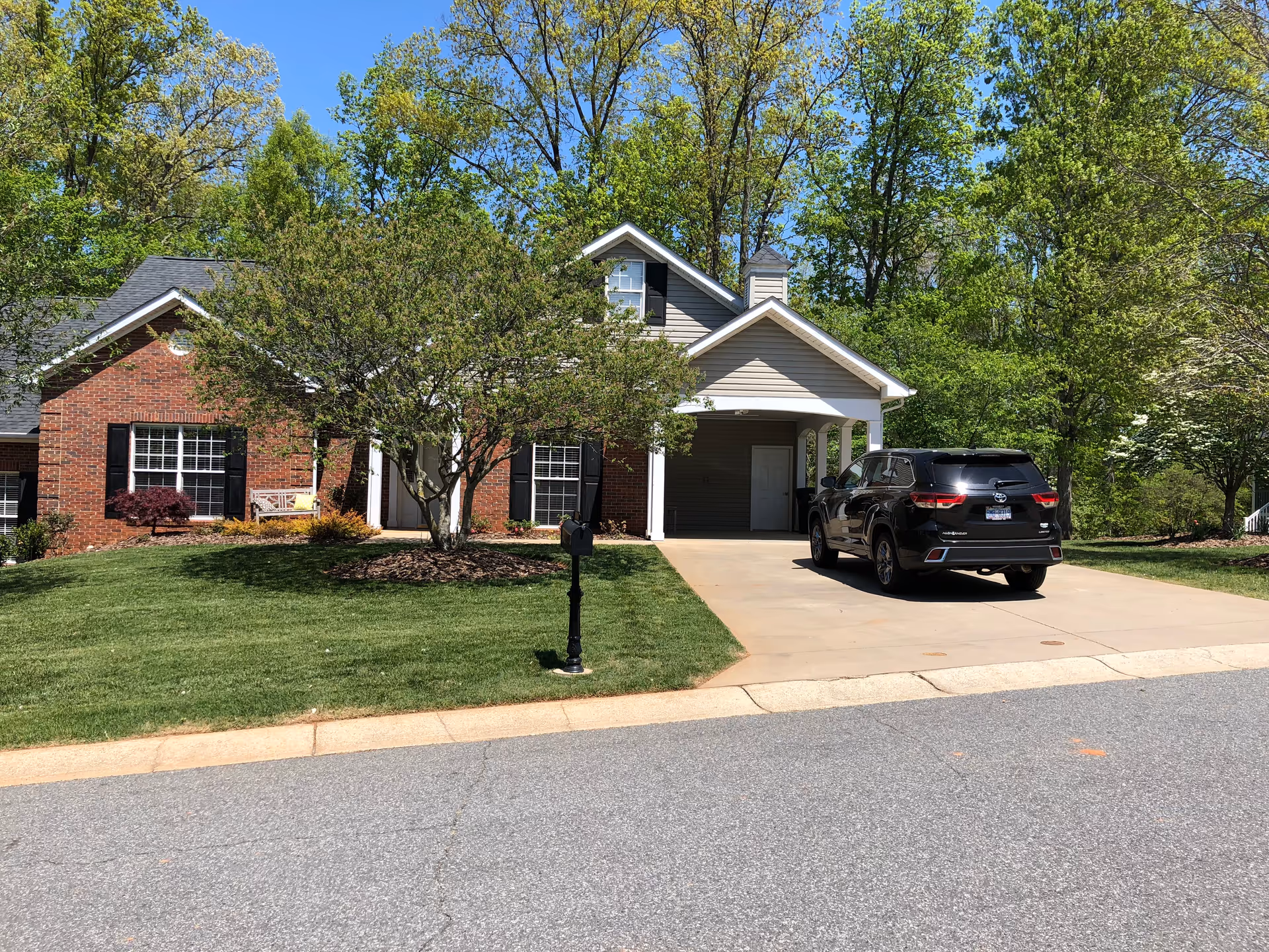 A suburban house with a brick and siding exterior, a tree in the front yard, a black SUV parked in the driveway, and green grass and trees surrounding the property under a clear blue sky.