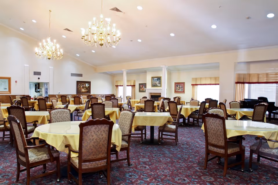 A spacious dining room with multiple round and rectangular tables covered with yellow tablecloths. Each table is surrounded by wooden chairs with patterned upholstery. The room features chandeliers hanging from the ceiling, large windows with beige curtains, framed artwork on the walls, and a piano in the corner.