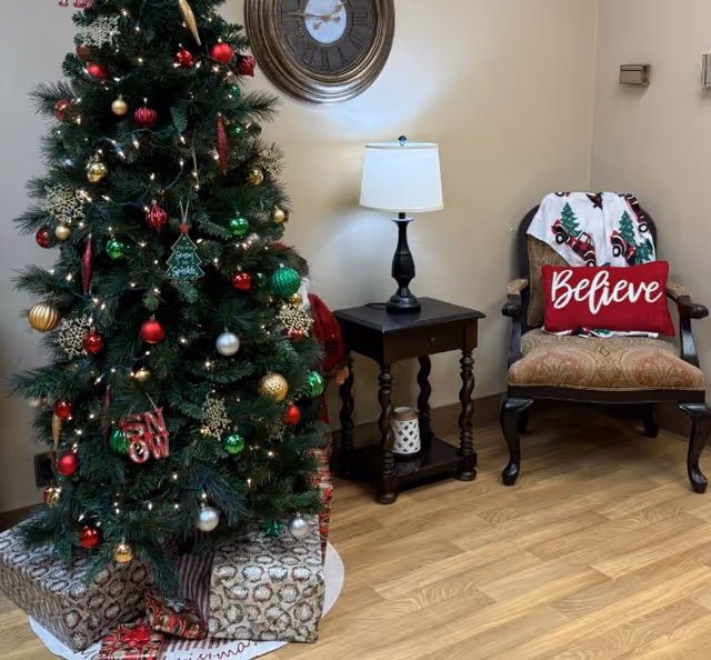 A decorated Christmas tree with red, green, gold, and silver ornaments and wrapped presents underneath. Next to the tree is a wooden side table with a lamp and a decorative candle. To the right is an upholstered armchair with a red pillow that says 'Believe' and a holiday-themed blanket draped over the back. A large round wall clock hangs on the beige wall behind the table.