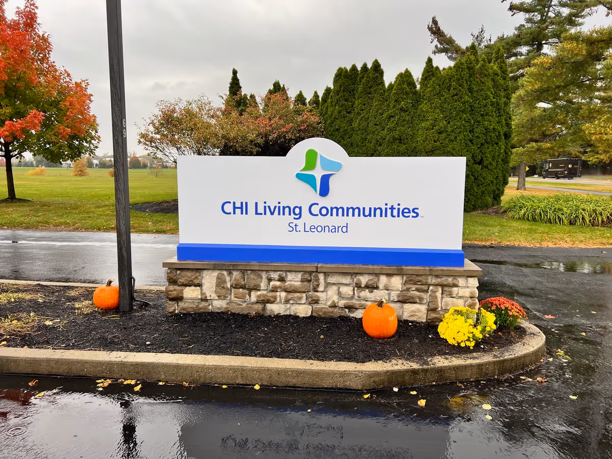 Entrance sign reading 'CHI Living Communities St. Leonard' on a stone base with pumpkins and fall landscaping in front of wet pavement.