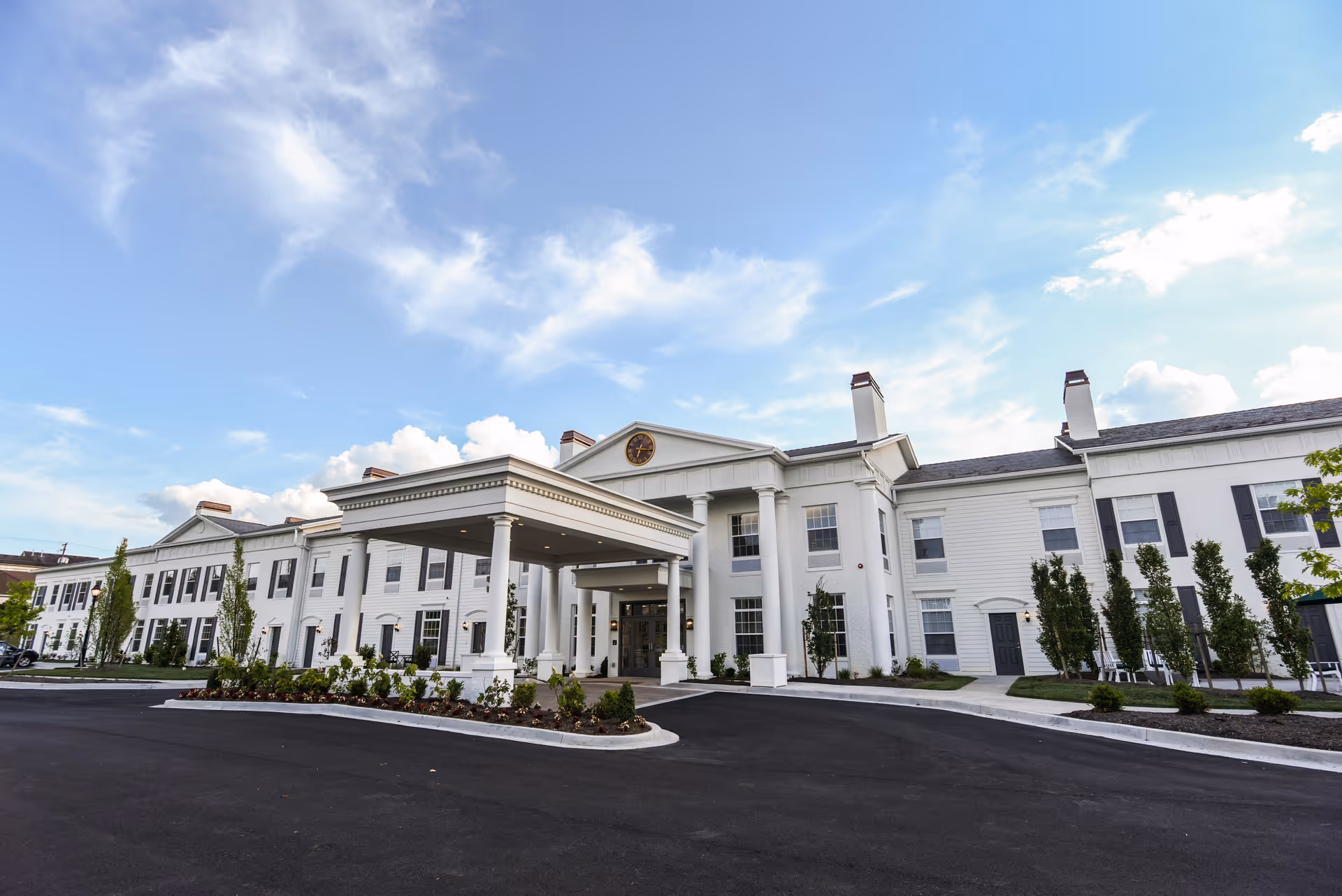 Front exterior view of a large, white senior living facility building with columns at the entrance, a covered driveway, and neatly landscaped greenery under a partly cloudy blue sky.