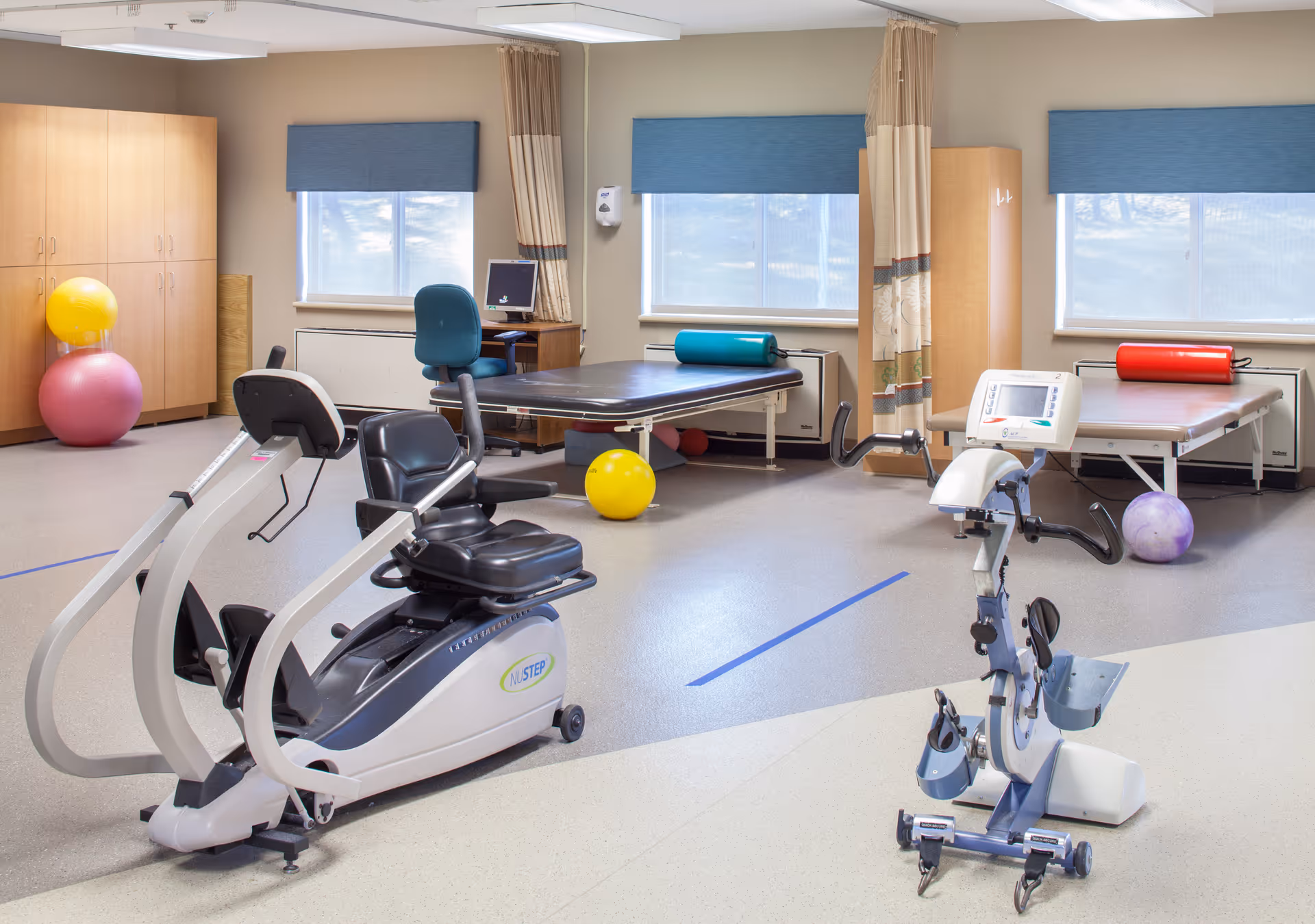 A rehabilitation or physical therapy room with exercise equipment including a recumbent exercise bike and a motorized pedal exerciser. The room has two therapy tables with cylindrical bolster pillows, colorful exercise balls, a desk with a computer, and large windows with blue blinds letting in natural light.