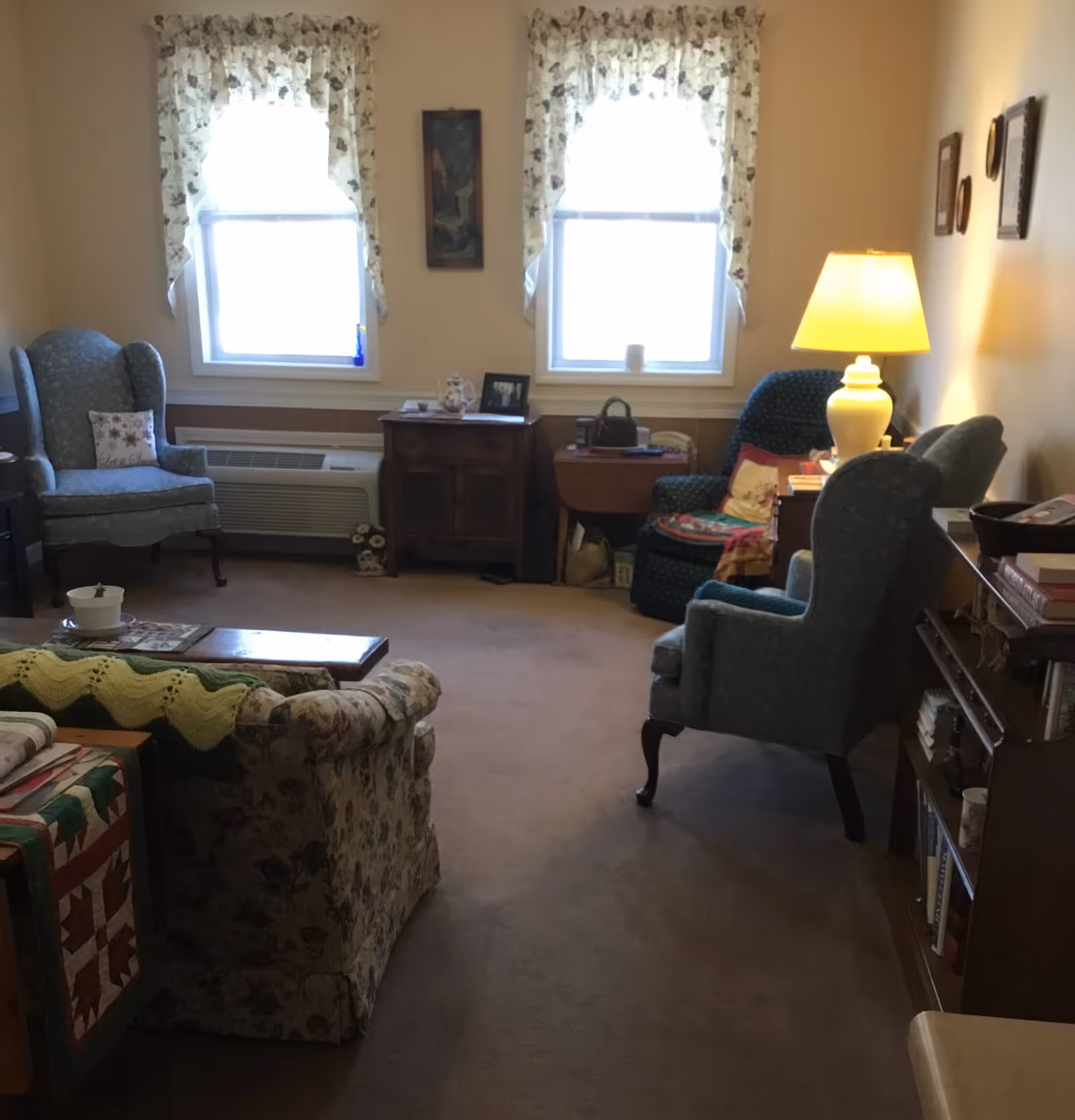 A cozy living room with two windows covered by floral curtains letting in natural light. The room contains a floral patterned sofa with a green and yellow knitted blanket draped over it, two upholstered armchairs, a wooden coffee table, a side table with a lamp, and a bookshelf filled with books. The walls are decorated with framed pictures.