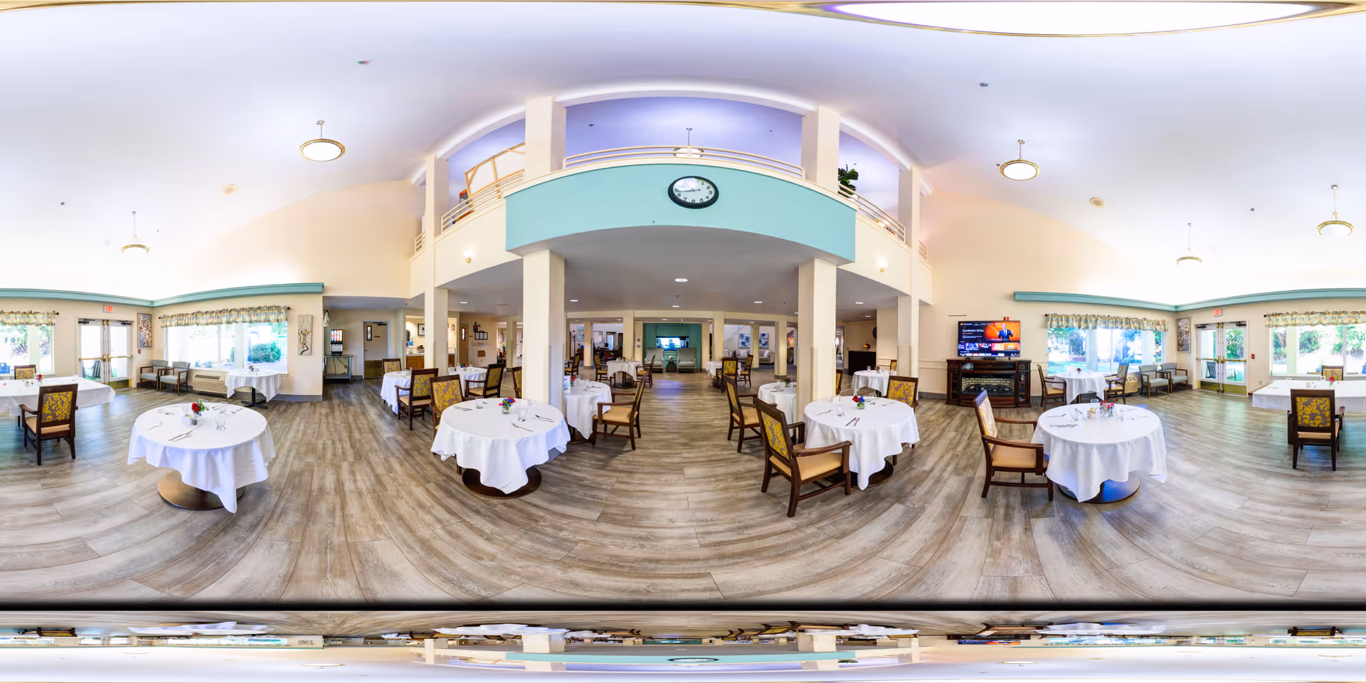 A spacious dining room with round tables covered in white tablecloths, each set with glassware and a small flower centerpiece. The room features wooden flooring, large windows with floral curtains allowing natural light, and a high ceiling with a mezzanine balcony above. Chairs with patterned upholstery are arranged around the tables, and a TV is mounted on the wall in the background.