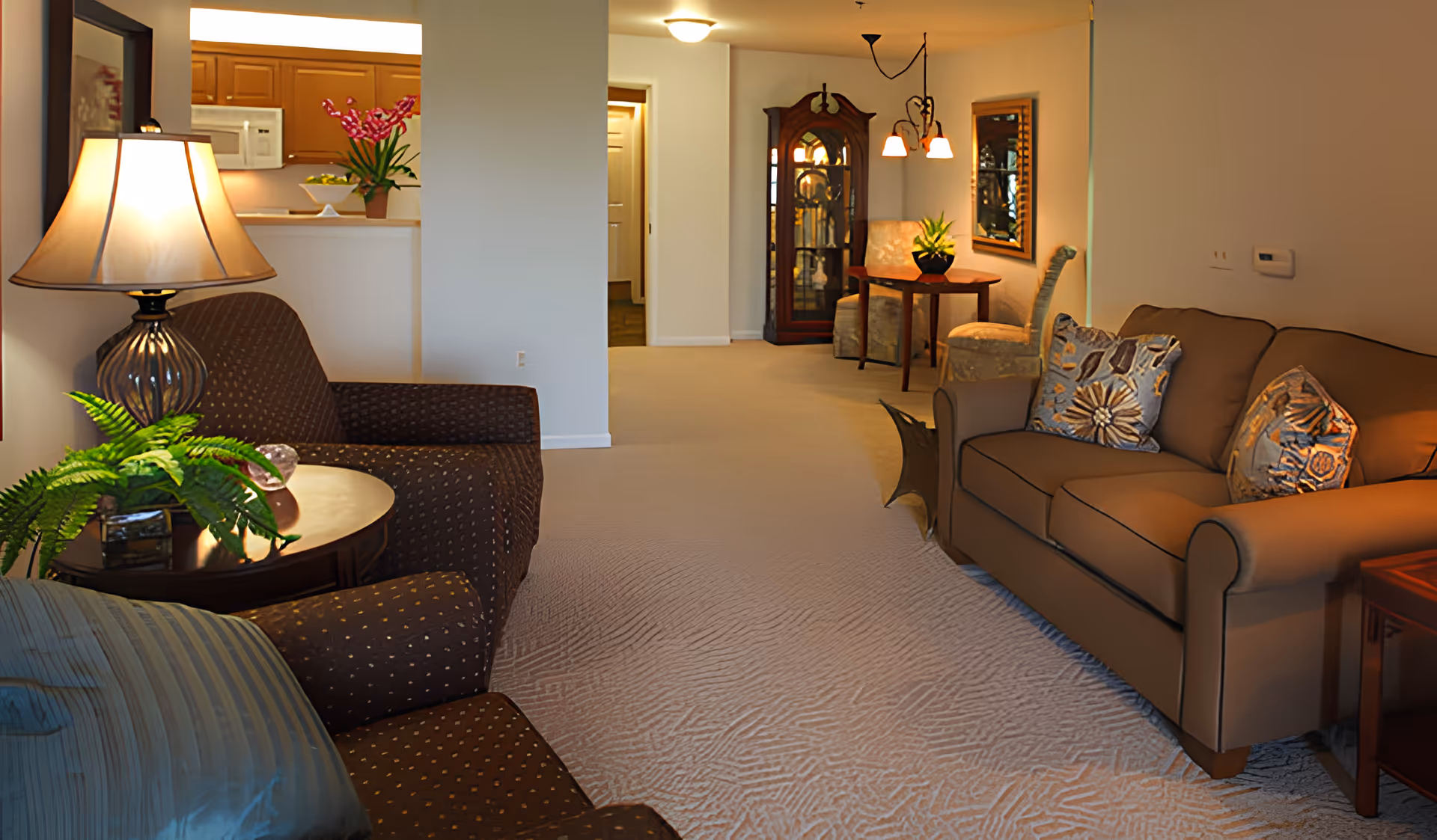A cozy living room area with a brown sofa adorned with patterned cushions on the right and a matching armchair with a striped pillow on the left. A round wooden side table with a lamp and a green plant is next to the armchair. In the background, there is a small dining area with a wooden table, two upholstered chairs, a decorative cabinet, and a mirror on the wall. The kitchen area with wooden cabinets and a microwave is partially visible through an open wall space.