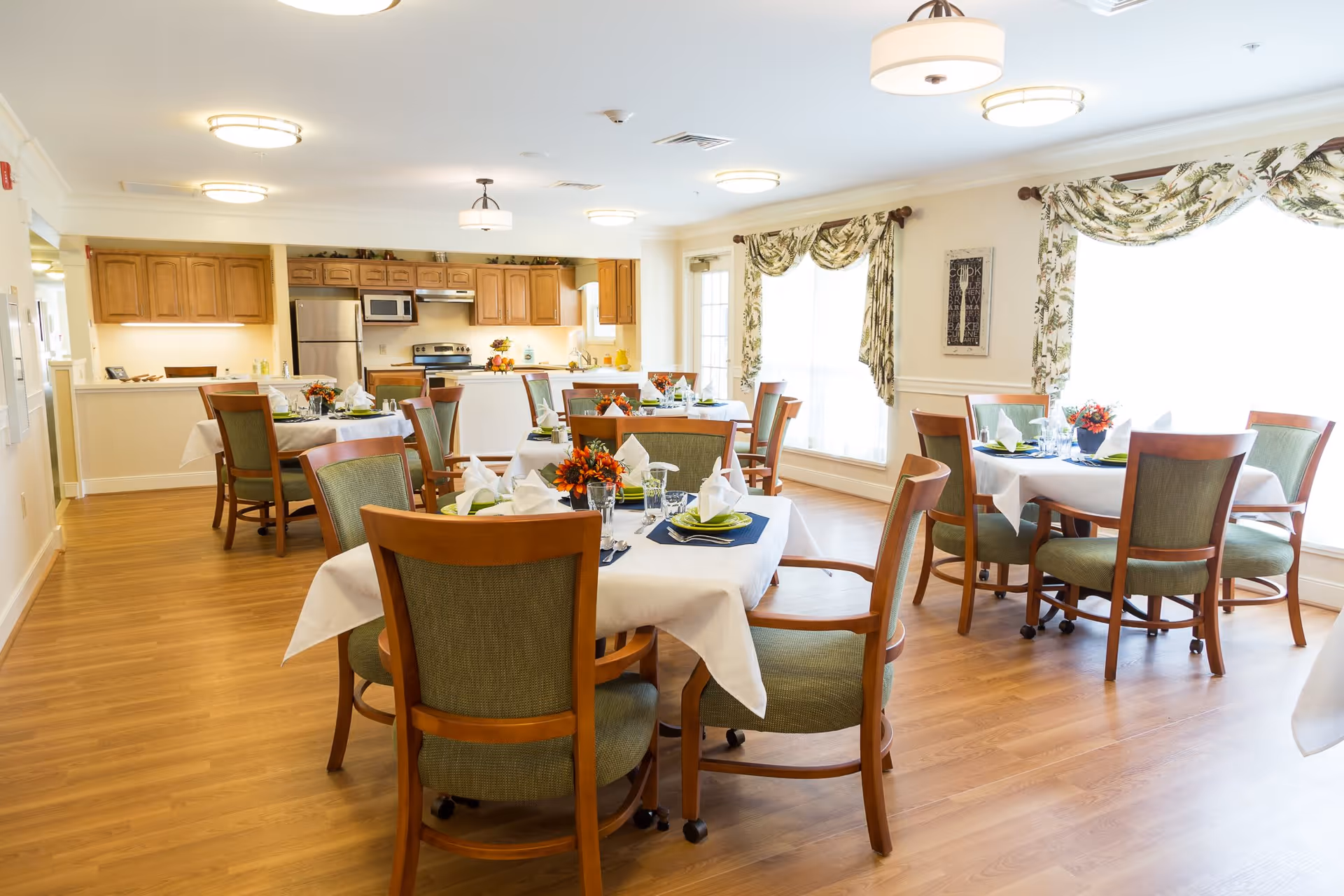 Bright dining room with several round tables covered with white tablecloths, each set with napkins, plates, glasses, and floral centerpieces. The room has wooden floors, green cushioned wooden chairs, large windows with floral curtains, and a kitchen area with wooden cabinets and stainless steel appliances in the background.