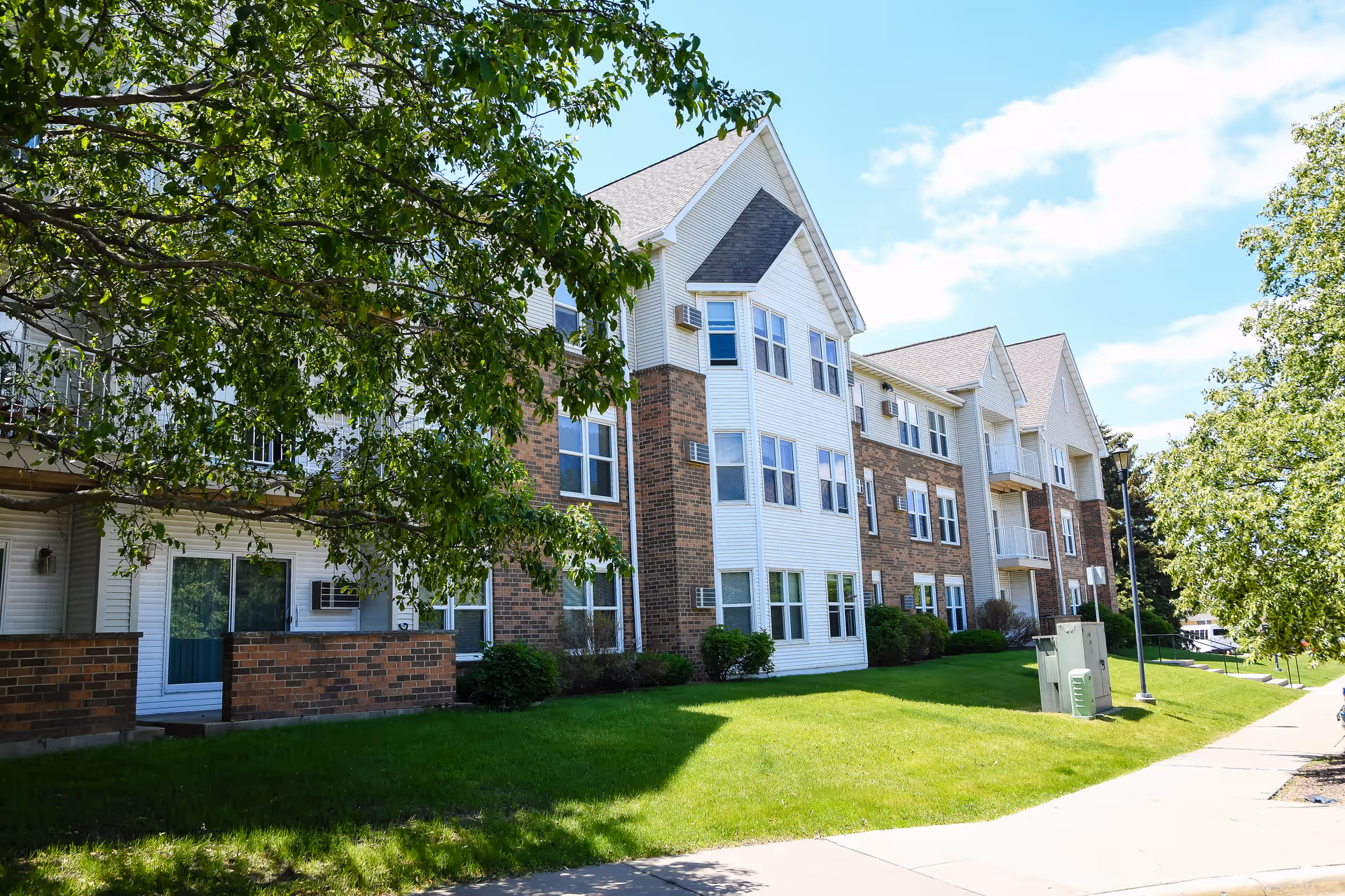 Three-story brick-and white-siding apartment building with balconies and a green lawn in front under a partly cloudy sky.