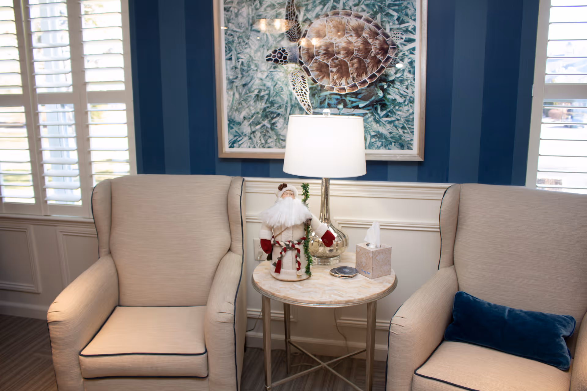 Two beige upholstered armchairs with dark piping are positioned on either side of a round marble-top side table. On the table is a lamp with a white shade, a small Santa Claus figurine, a tissue box, and a small dish. Behind the table is a wall with blue vertical stripes and a framed picture of a sea turtle. White plantation shutters cover the windows on either side of the seating area.