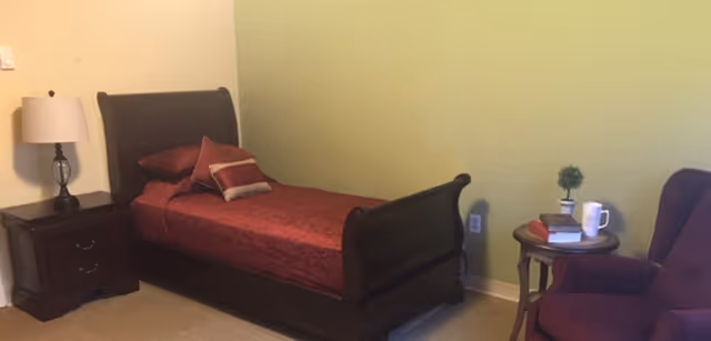 A bedroom with a dark wooden sleigh bed dressed in red bedding and pillows. Next to the bed is a dark wooden nightstand with a lamp on it. There is a small round side table with a potted plant, a book, and a white mug. A purple upholstered armchair is positioned near the side table. The walls are painted light green and beige.