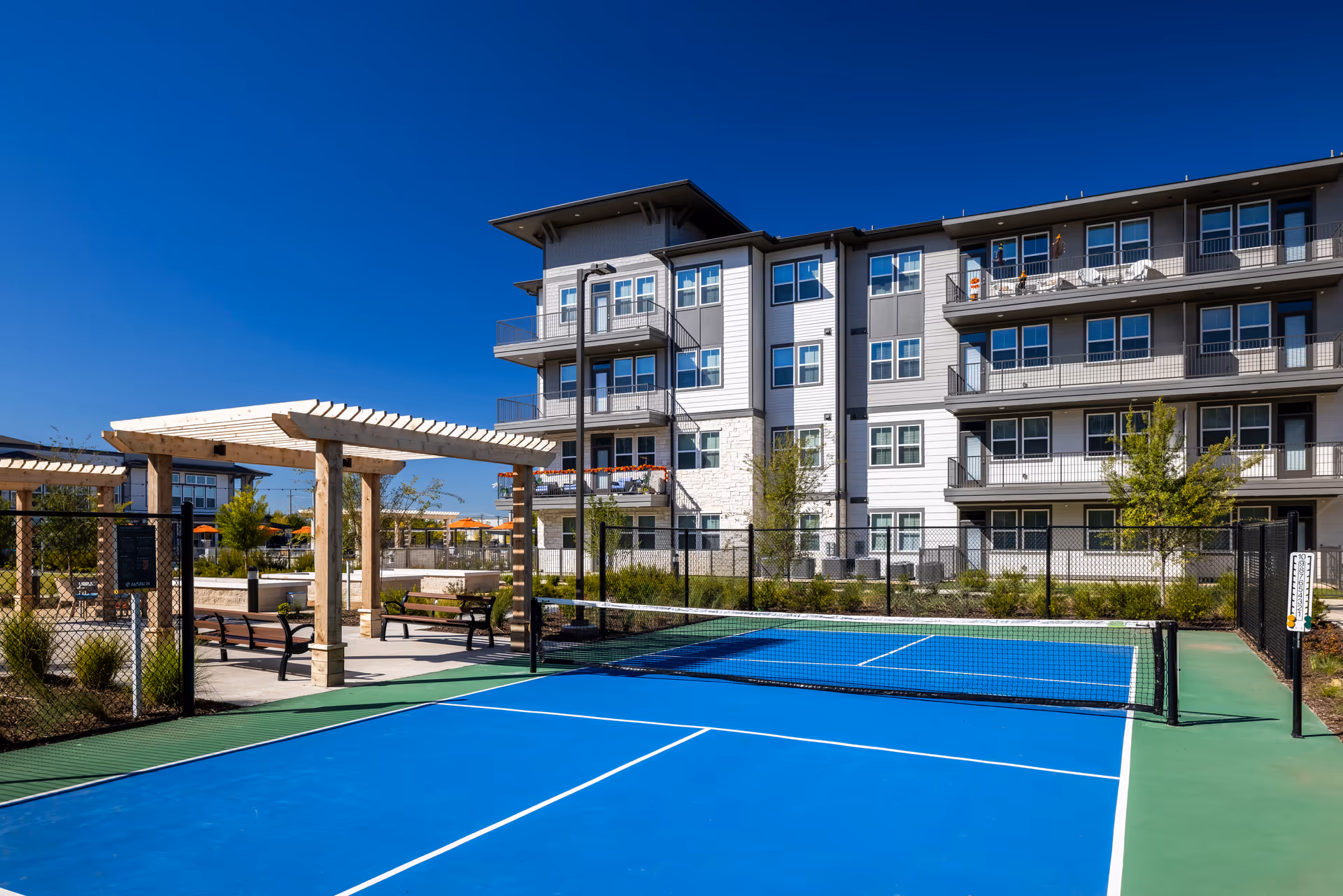 View of the exterior of Amberlin at The Station featuring a blue tennis court, pergola, and landscaped area with benches.