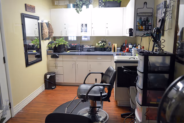 Interior of a small salon or grooming room with a black salon chair in the center, white cabinets along the back wall, a mirror on the left wall, and various hair styling tools and products on the counter and shelves.
