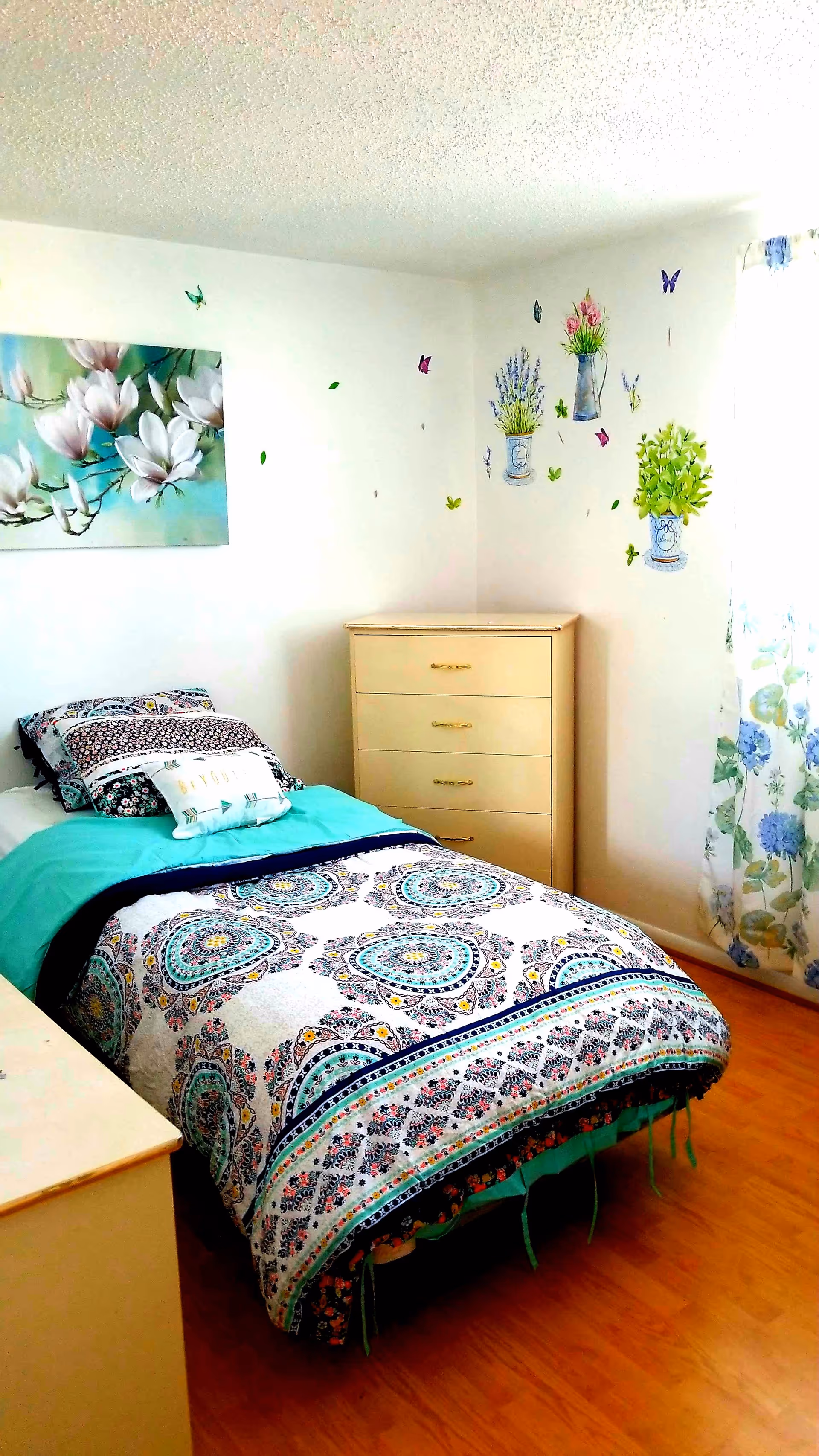 A small bedroom with a single bed covered in a colorful patterned bedspread and pillows. There is a light wood dresser with four drawers in the corner. The walls are decorated with floral and butterfly decals, and a painting of white flowers hangs above the bed. A window with floral curtains allows natural light into the room, which has a wooden floor.