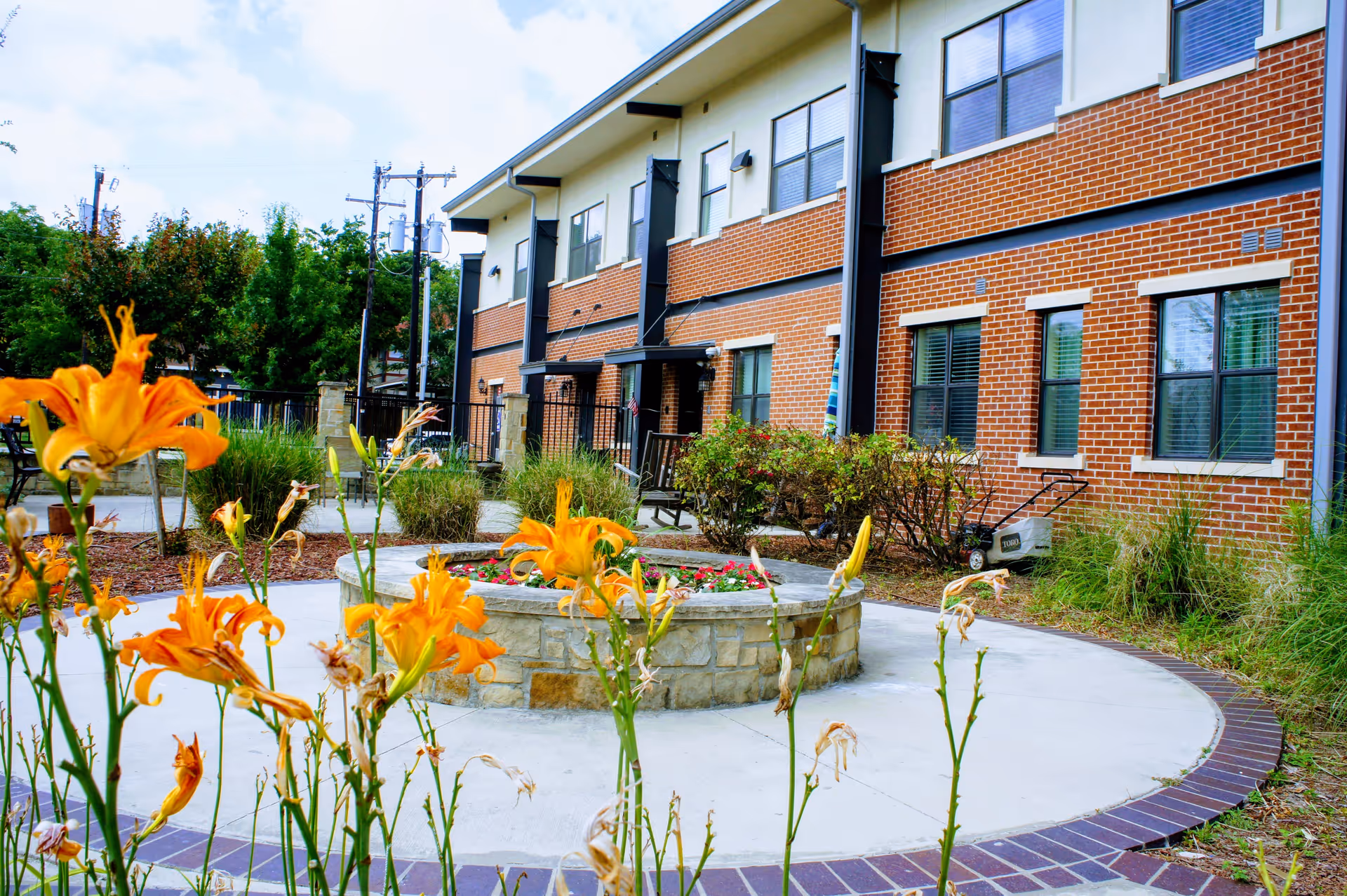 Outdoor courtyard with orange flowers and a circular stone planter in front of a two-story brick senior living building.