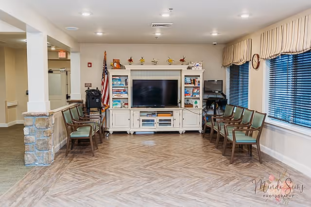 A spacious common area with a large TV on a white entertainment center filled with books and decorative items. There are two rows of green cushioned chairs facing the TV, an American flag to the left, and large windows with striped valances on the right. The floor has a wood-patterned design and the walls are painted light beige.