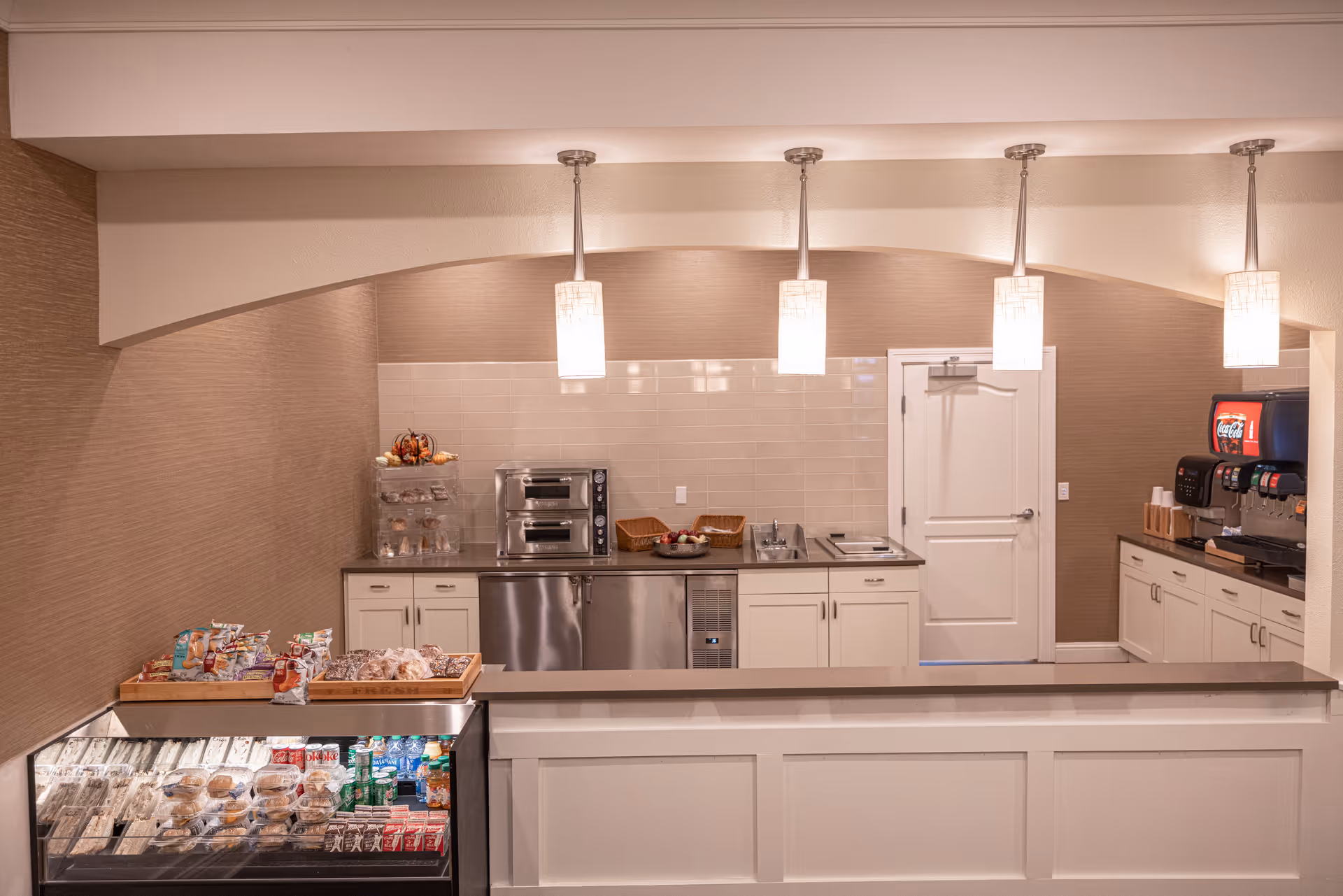 A small kitchen or snack bar area with a counter in the foreground. Behind the counter are cabinets, a small oven, a sink, a refrigerated display case with sandwiches and drinks, and a soda fountain machine. The area is well-lit with four hanging pendant lights and has a neutral color scheme with beige walls and white cabinetry.