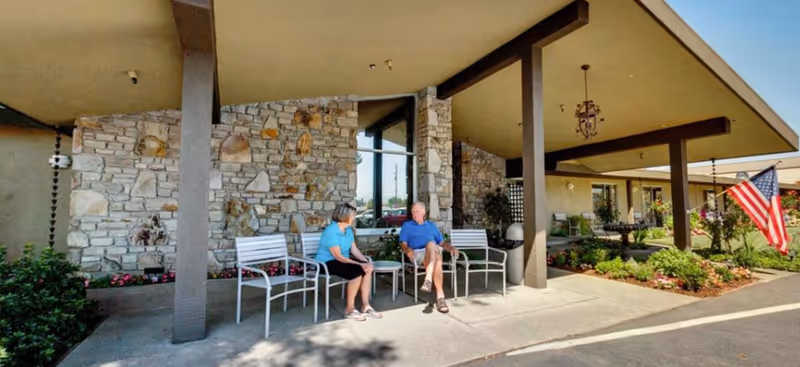 Two elderly people sitting and talking on white benches under a covered patio area outside a stone and beige building with plants and an American flag nearby.