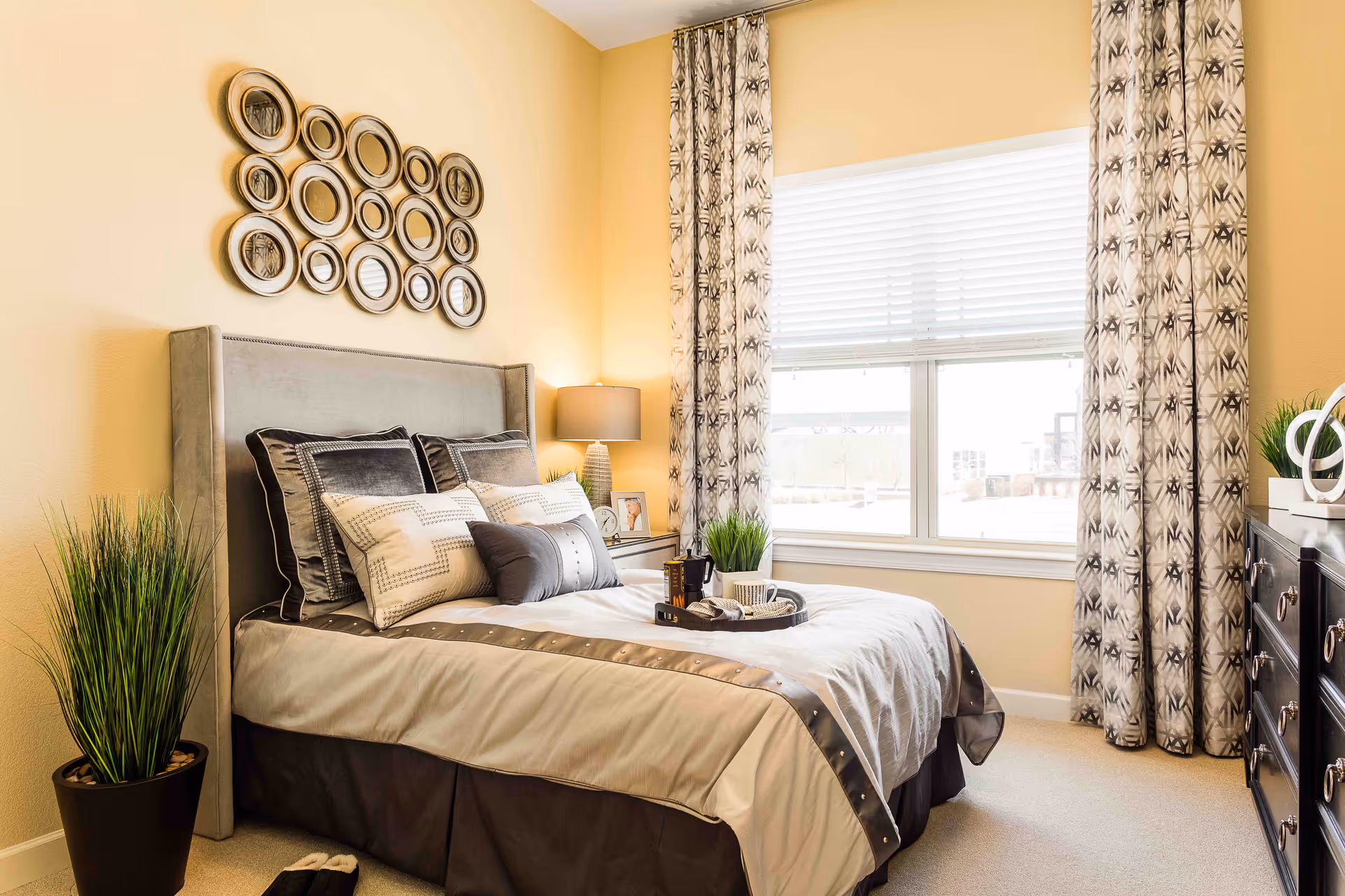 A cozy bedroom with a neatly made bed featuring multiple pillows and a tray with a cup and book. The room has a large window with patterned curtains, a bedside table with a lamp and picture frame, a dresser with decorative items, and a potted plant in the corner.
