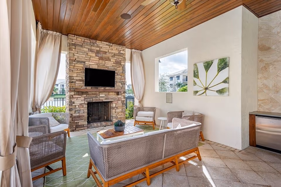 A covered lounge with wicker sofas and chairs facing a stone fireplace and wall-mounted TV under a wood-paneled ceiling.