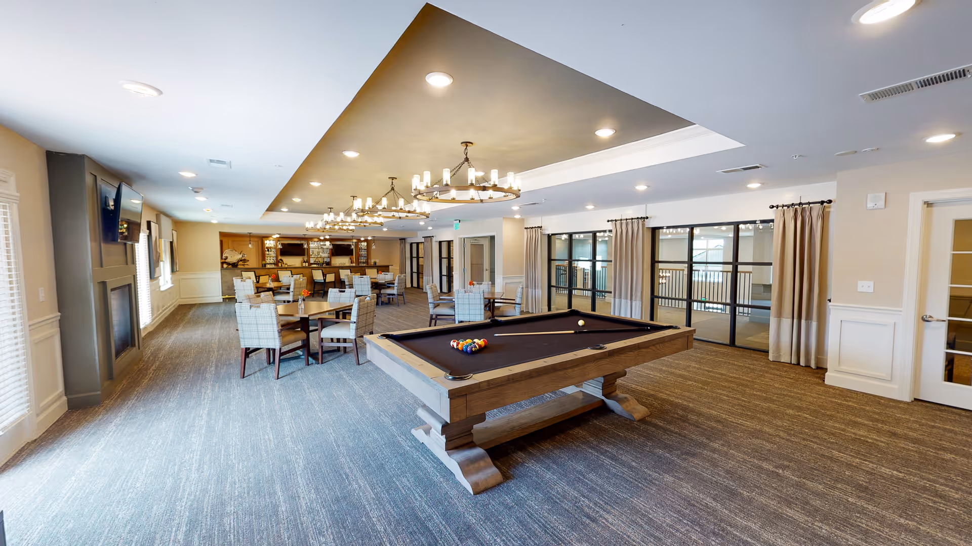 A spacious and well-lit common area featuring a pool table in the foreground, several round tables with cushioned chairs, a fireplace on the left wall, and a bar area with stools in the background. The room has large windows with curtains, modern chandeliers, and recessed ceiling lights.