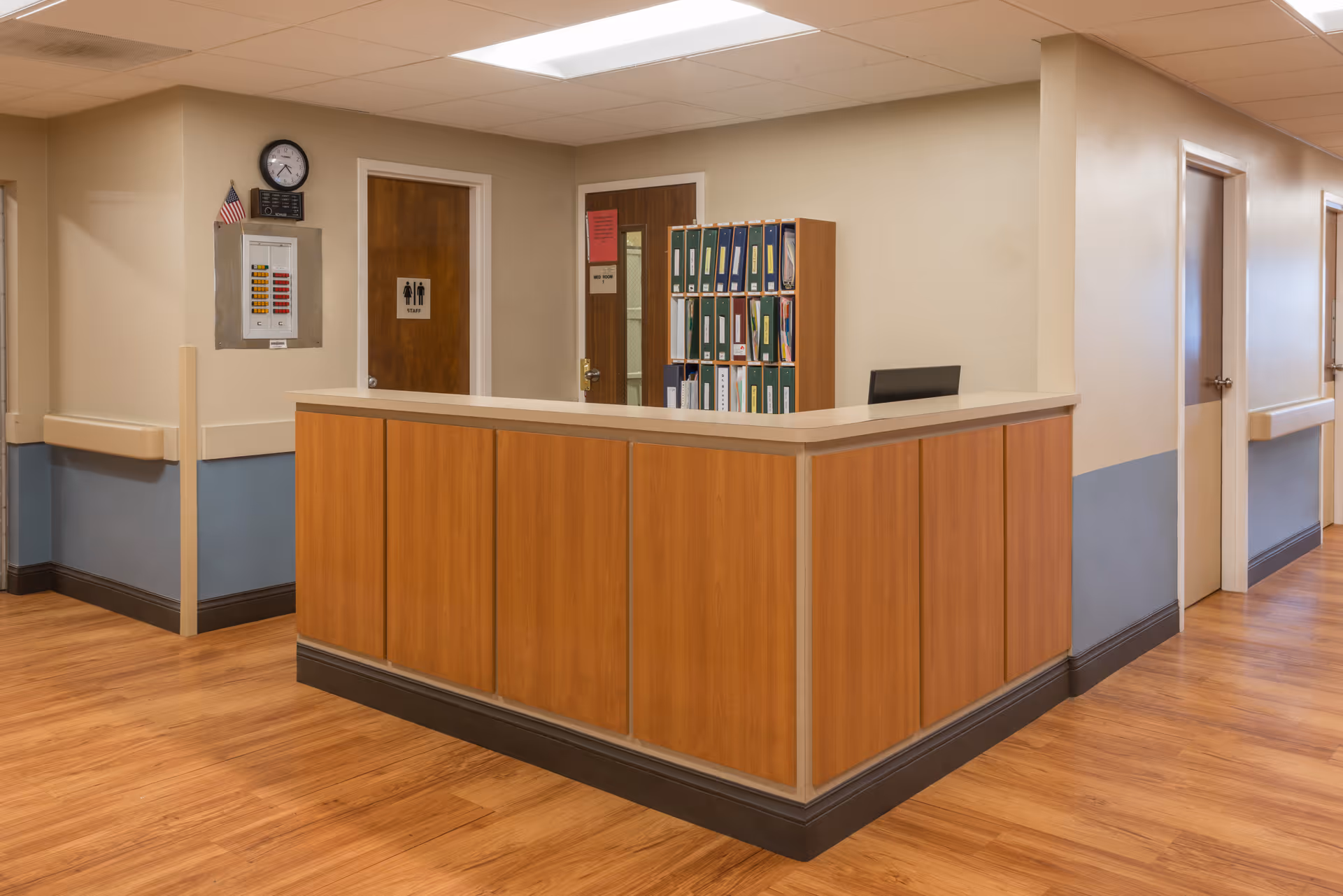 Reception desk area in a healthcare facility with wood paneling, a clock on the wall, an American flag, a fire alarm panel, and several doors including one marked as staff restroom. There are shelves with organized binders behind the desk.