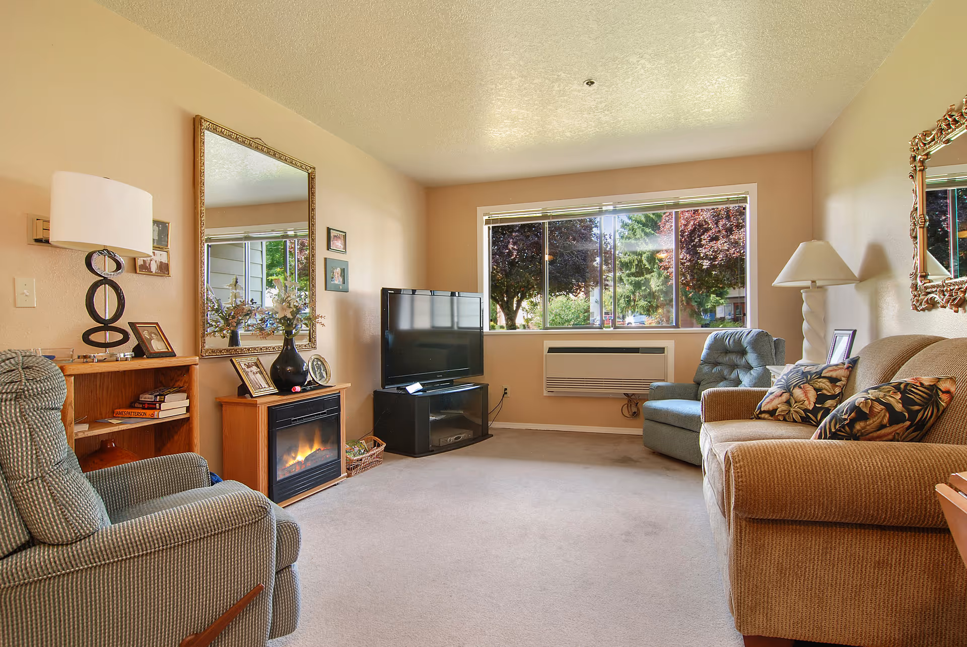 A cozy living room with beige walls and carpet, featuring a brown sofa with floral cushions, two green armchairs, a flat-screen TV on a black stand, an electric fireplace with a wooden mantel, a large mirror above the fireplace, a wooden bookshelf with books and framed photos, a table lamp, and a large window showing trees outside.