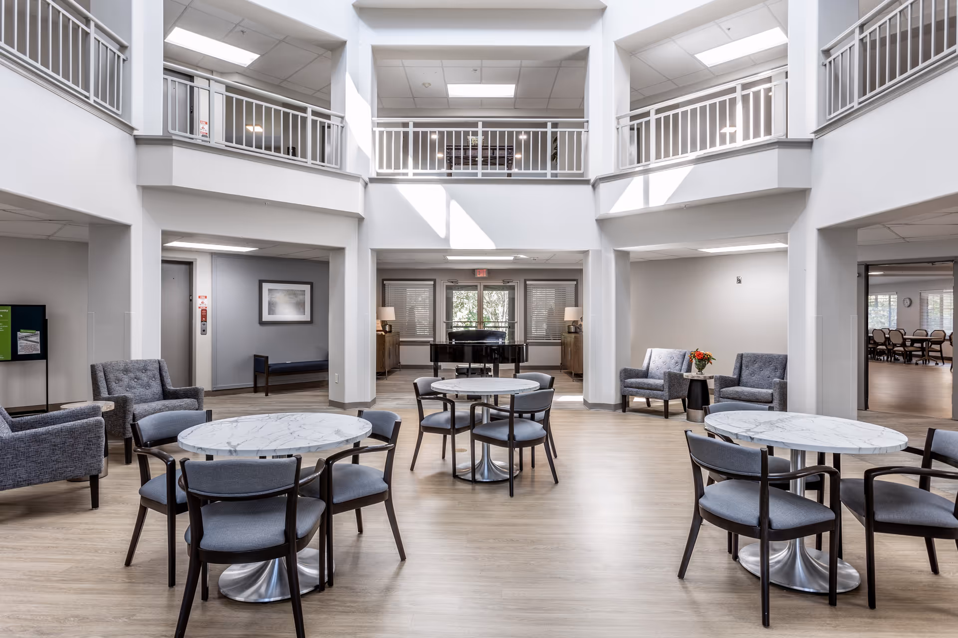 A bright and spacious common area in a senior living facility with round marble-top tables surrounded by chairs, several gray upholstered armchairs, and a grand piano in the background. The room features high ceilings with a second-floor balcony railing and large windows letting in natural light.
