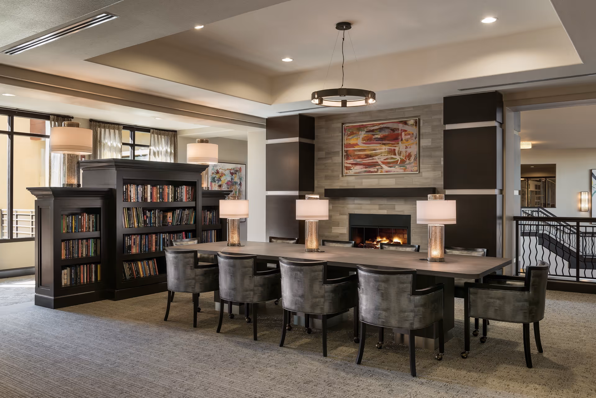 A cozy and modern common area featuring a long wooden table surrounded by eight gray upholstered chairs. The table is decorated with four lamps with beige shades. Behind the table is a built-in fireplace with a colorful abstract painting above it. To the left, there are large windows with sheer curtains and a dark wood bookshelf filled with books. The room has a neutral color palette with soft lighting and a carpeted floor.