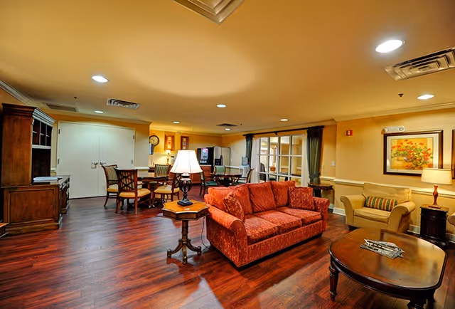Well-lit communal living room with a red sofa, armchairs, coffee and side tables, a dining table and entertainment cabinet on hardwood floors.