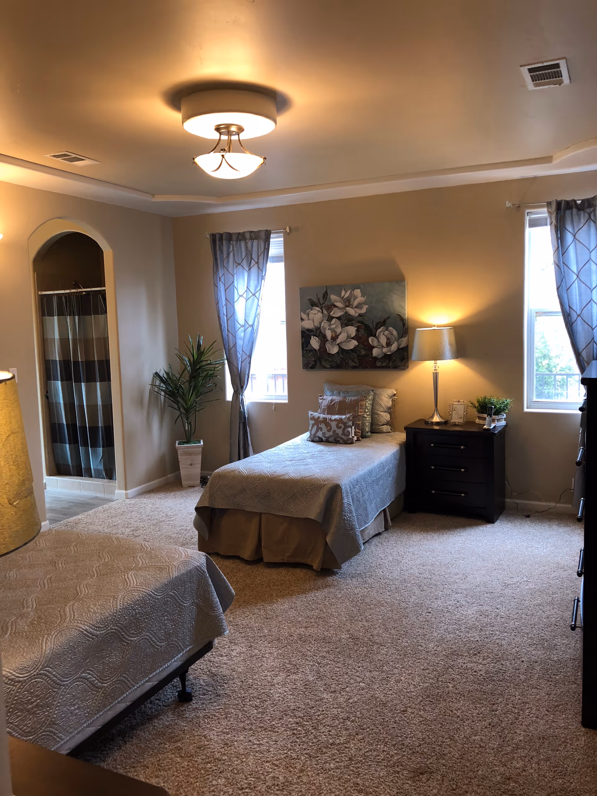 A cozy bedroom with two twin beds covered in light gray bedspreads. There is a nightstand with a lamp, a small plant, and a picture frame between the beds. Two windows with patterned curtains allow natural light into the room. A floral painting hangs on the wall above one bed. In the corner, there is a potted plant next to an open doorway leading to a bathroom with a striped shower curtain. The room has beige walls and carpeted flooring.
