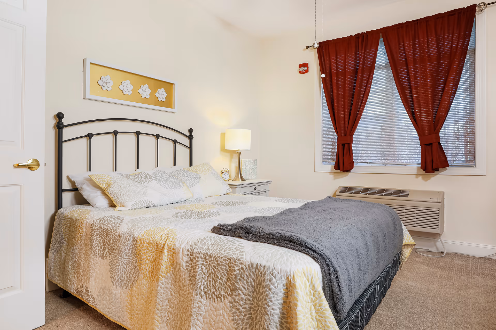 A bedroom with a bed featuring a black metal headboard, beige and white patterned bedding, and a gray blanket folded at the foot. There is a white nightstand with a lamp and a clock beside the bed. A window with red curtains and blinds is on the right wall, and an air conditioning unit is installed below the window.