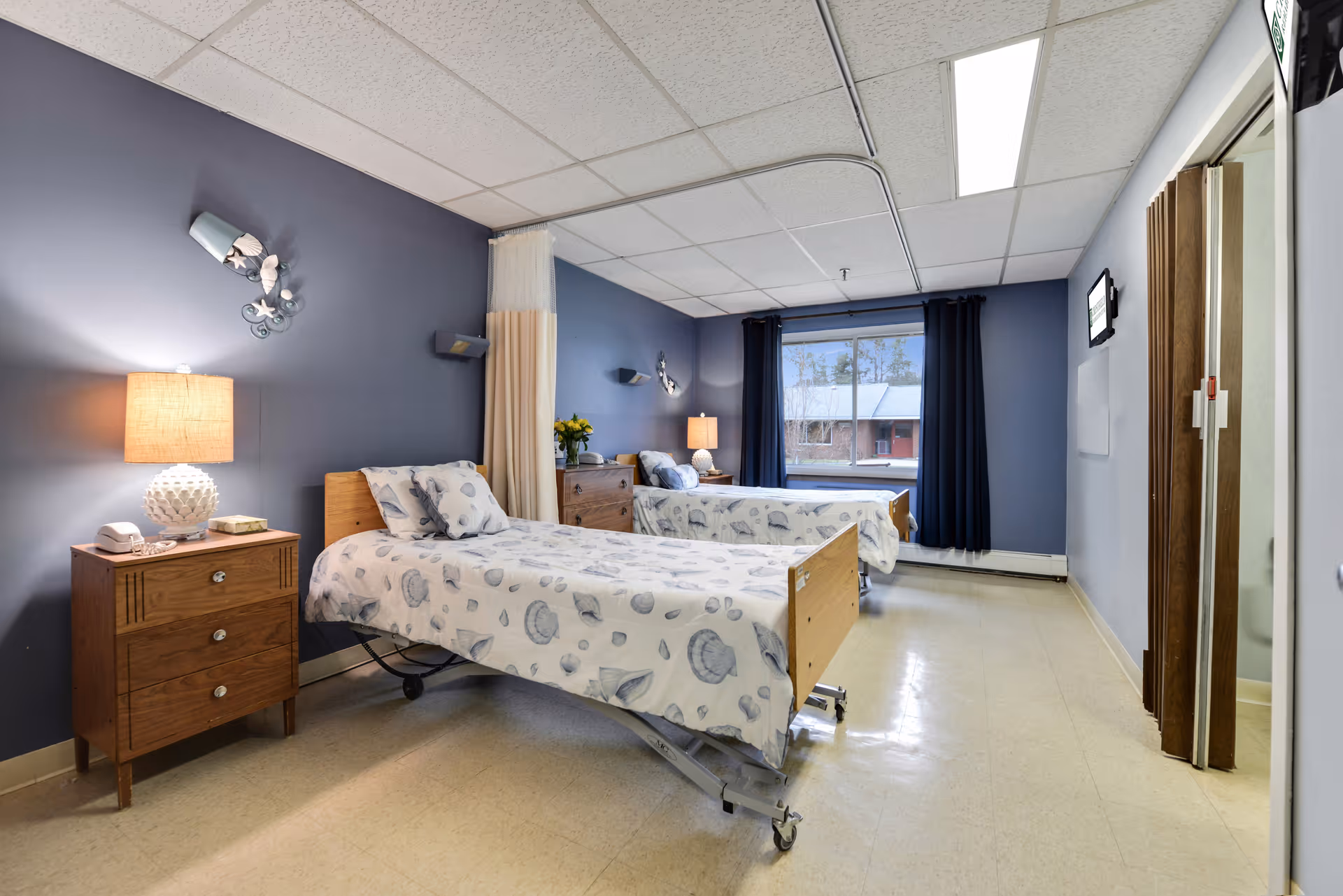 A shared patient bedroom with two adjustable hospital beds, nightstands, lamps, and a large window with dark curtains.