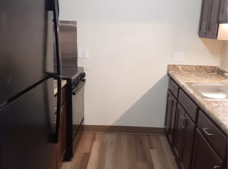 Small galley kitchen with a black refrigerator and stove on the left and a sink with dark wood cabinets and laminate countertops on the right.