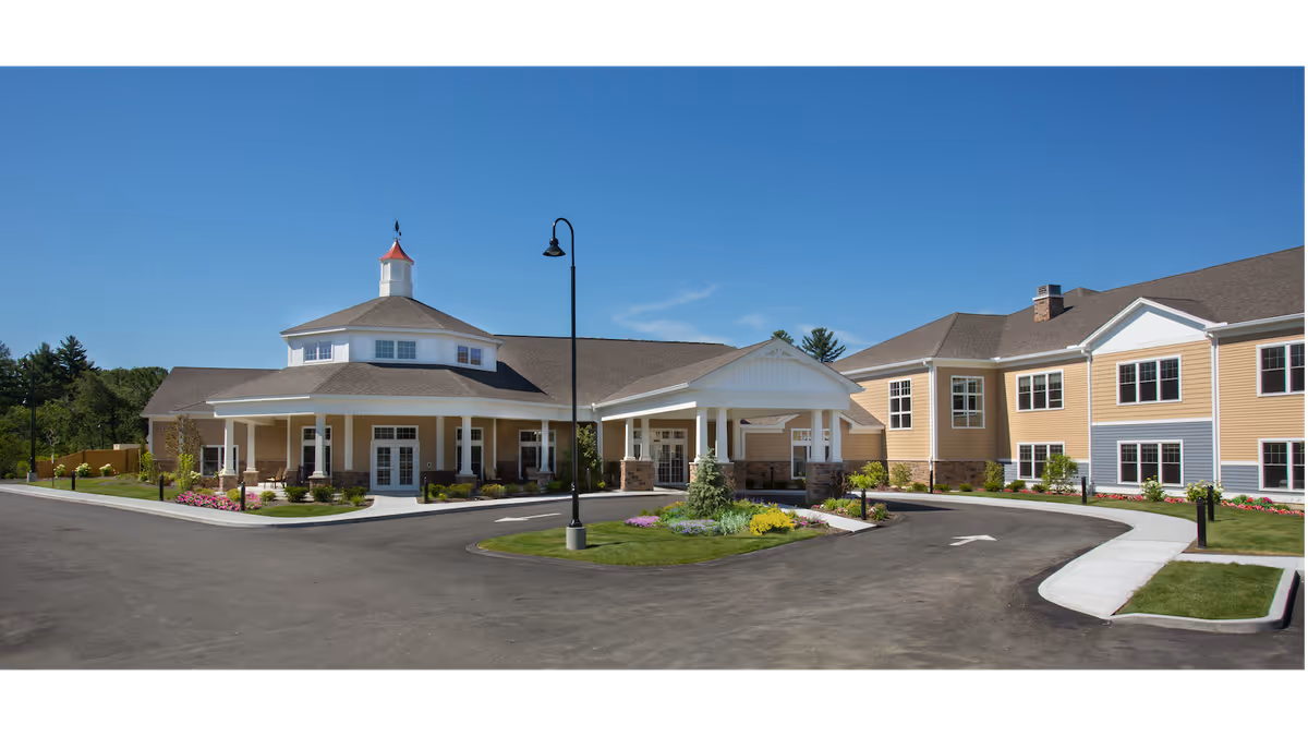 Exterior view of a large, modern assisted living facility building with beige siding and white trim. The building has multiple windows, a covered entrance with columns, a circular driveway, landscaped greenery, and a clear blue sky overhead.