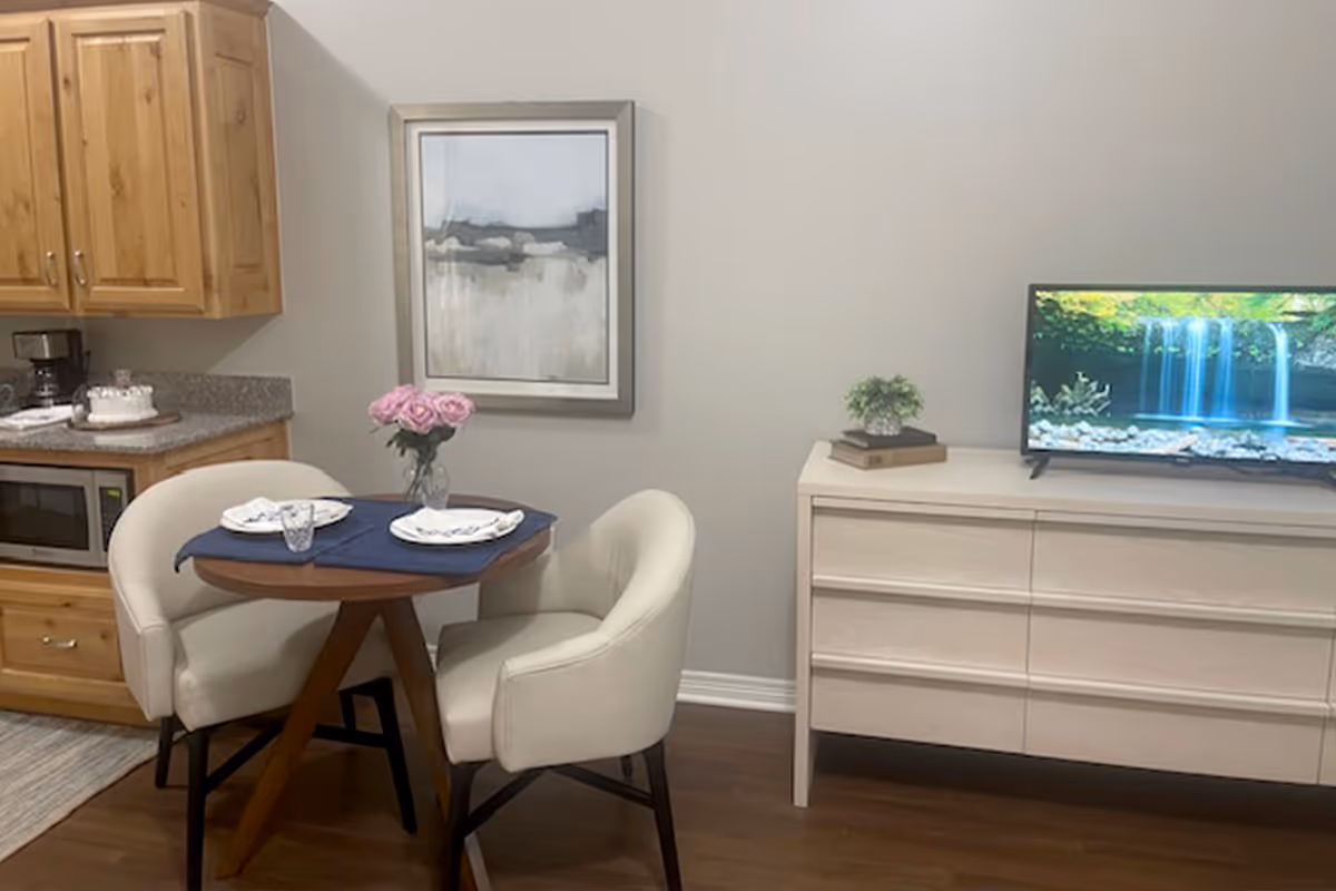 A cozy dining area with a small round wooden table set for two with plates, glasses, and a vase of pink roses. Two cream-colored upholstered chairs surround the table. To the left, there is a kitchen counter with wooden cabinets, a microwave, and a coffee maker. On the right, a light-colored dresser holds a small plant and a TV displaying a waterfall scene. A framed abstract painting hangs on the wall above the table.