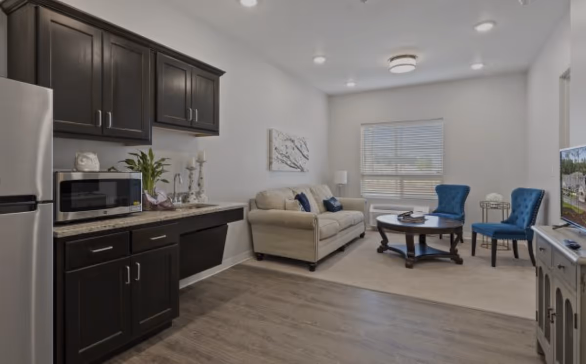 A modern living area with a small kitchen on the left featuring dark cabinets, a microwave, and a refrigerator. The living room has a beige sofa with blue pillows, two blue upholstered chairs, a round wooden coffee table, a window with blinds, and a TV on a stand to the right.
