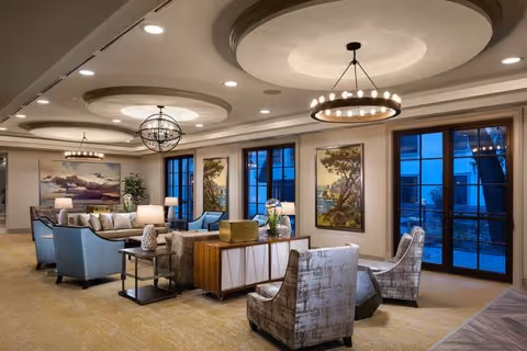 A spacious and elegantly furnished living room with multiple seating areas including blue and patterned armchairs, sofas, and side tables with lamps. The room features large windows and glass doors, decorative ceiling lights, and framed landscape paintings on the walls.