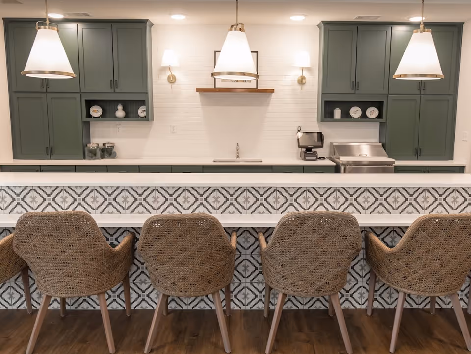 A modern communal kitchen/bar area with four woven barstools facing a patterned tile counter, green cabinets, and pendant lights.