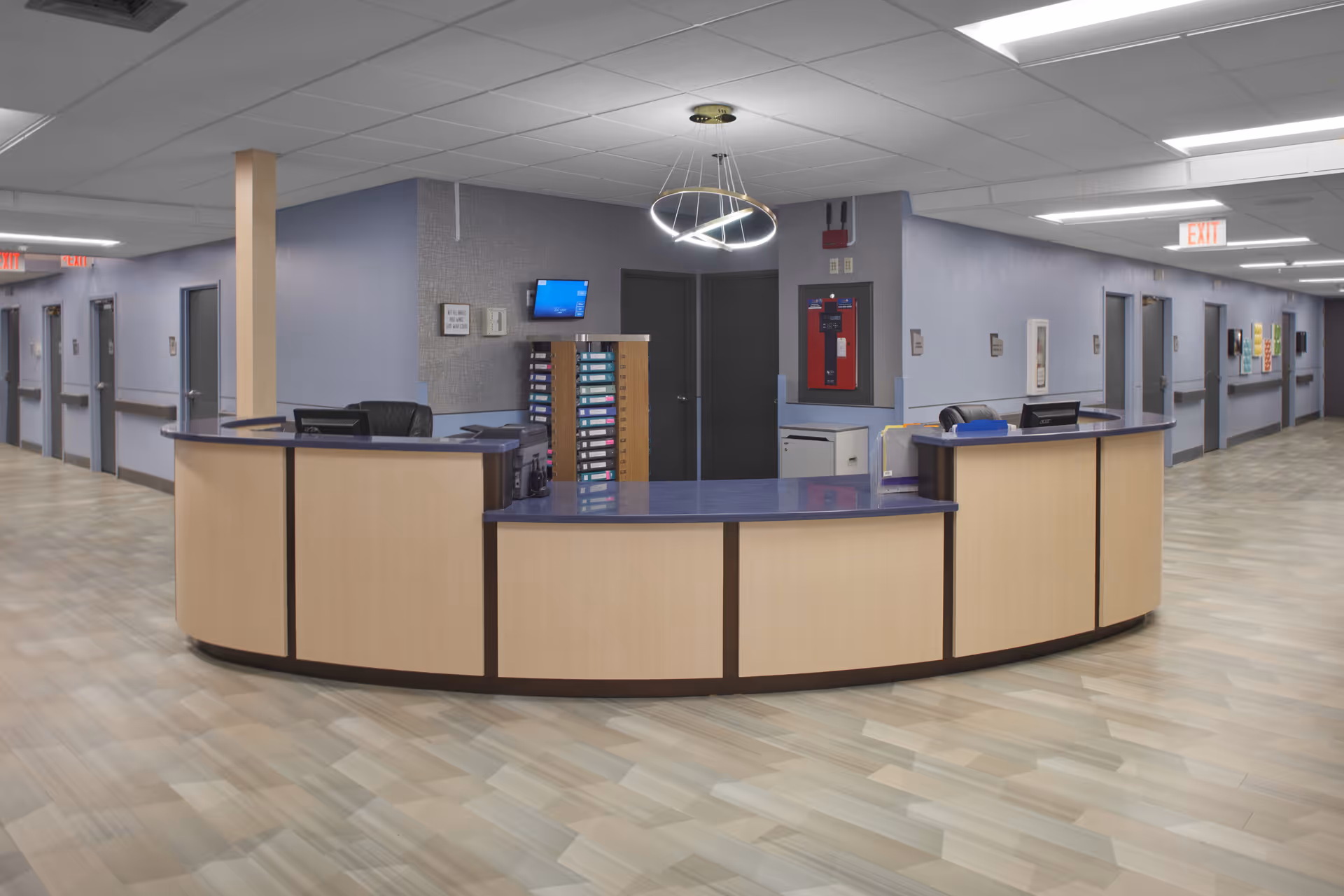 Curved reception/nurses' station in the center of a wide interior hallway with doors, exit signs, and a pendant light overhead.