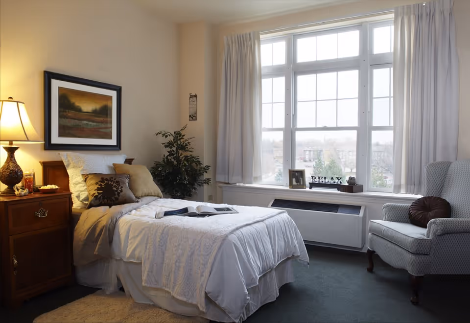 A cozy bedroom with a single bed made with white and beige bedding, a wooden nightstand with a lamp and decorative items, a framed landscape painting on the wall, a potted plant in the corner, a large window with white curtains letting in natural light, and a cushioned armchair with a round pillow.