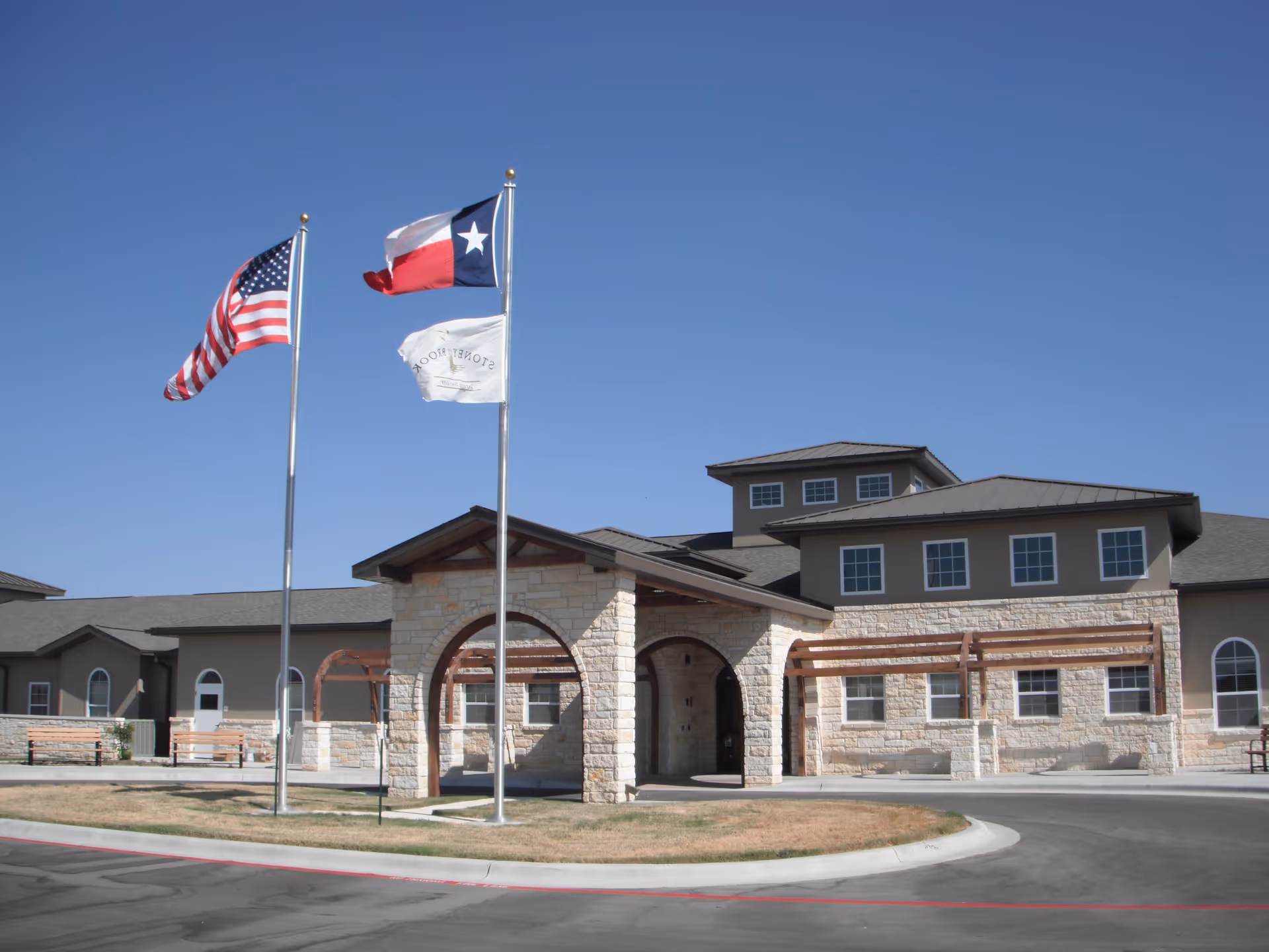 Exterior view of Stoney Brook of Hewitt facility with a stone and stucco facade, three flags on flagpoles including the American flag, Texas state flag, and a white flag with the facility's name, under a clear blue sky.
