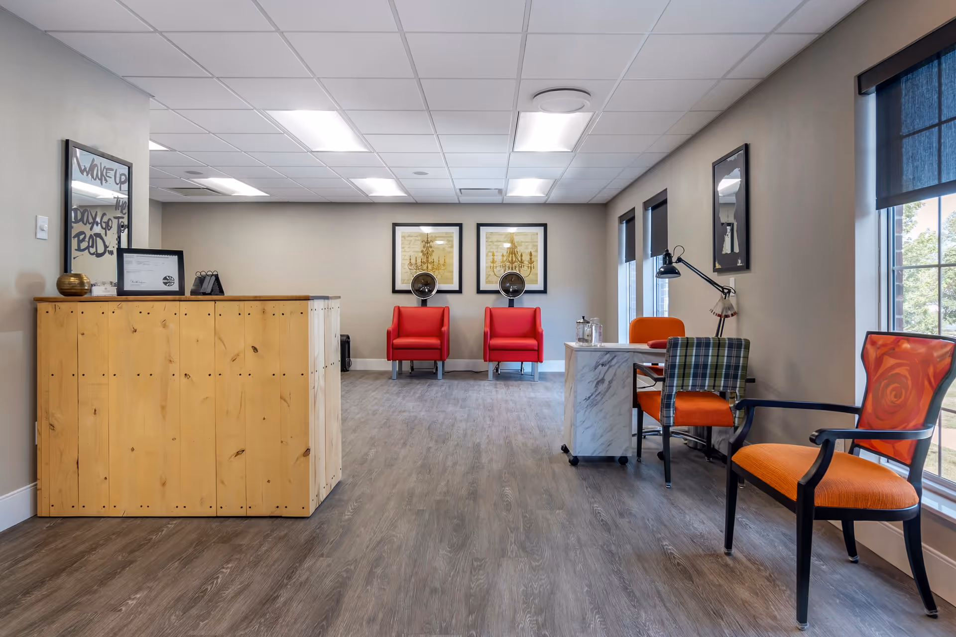 A bright and clean senior living community room with a wooden reception desk on the left, two red armchairs against the back wall with hair dryers above them, and a small desk with two orange chairs and a plaid chair on the right near large windows with black blinds.