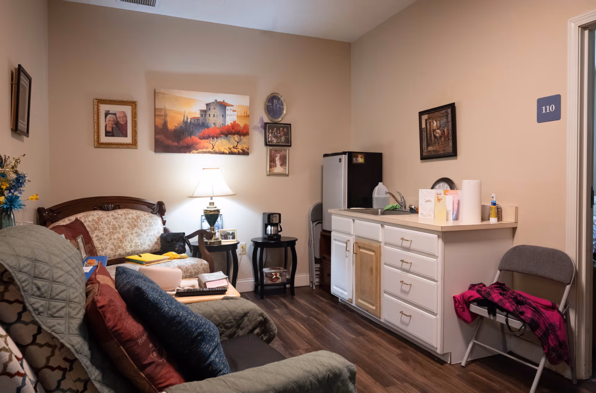 A cozy living area in a senior living facility, featuring a sofa, lamp, small kitchenette, and decorative artwork.